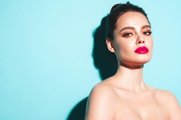 Fashion beauty portrait of young brunette woman with evening stylish makeup and perfect clean skin Sexy model with hair in a bun posing in studio near blue wall With pink bright natural lips