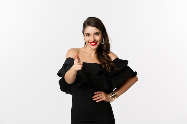 Fashion and beauty. Sassy pretty woman in black dress and makeup, pointing finger at camera to congratulate or praise, standing over white background