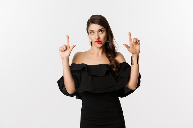 Fashion and beauty. Thoughtful attractive woman in black dress staring and pointing up, thinking with tempted look, white background