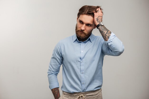 Fashion brutal hipster business man with tattoos in stylish elegant blue shirt fixing hair on a white background