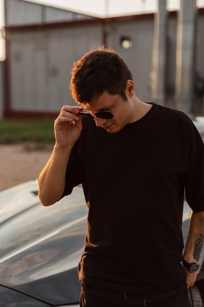 Fashion business man in black clothes walks near a car and puts on a stylish sunglasses at sunset