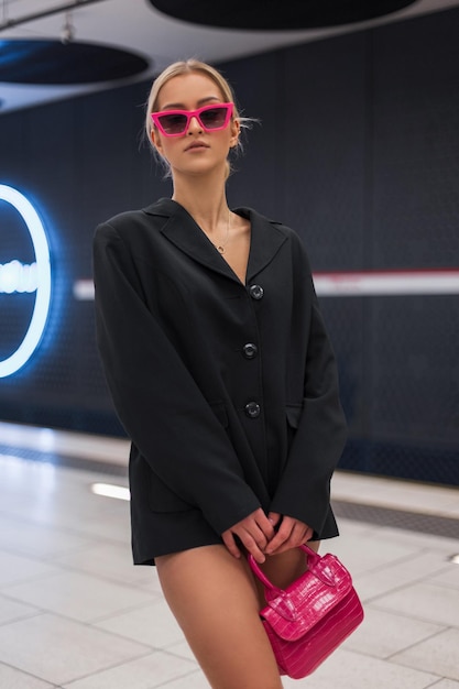 Fashion cool glamorous beautiful woman with pink sunglasses in a black blazer with a pink stylish handbag stands and poses at a white modern subway station