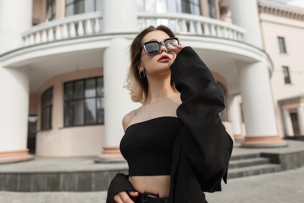 Fashion elegant pretty woman model in stylish trendy black clothes with top and blazer wears a cool modern sunglasses and walks in the city Urban female business style look outfit