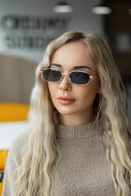 Fashion female portrait of a beautiful young woman with vintage sunglasses with a curly blond hairstyle in a stylish beige sweater sits in a cafe