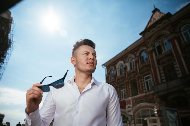 Fashion man in a white shirt is standing on the street at a sunny day is posing watching holding glasses