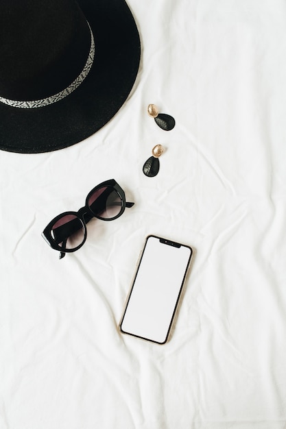 Fashion mockup composition with elegant womens accessories on white linen. Hat, sunglasses, earrings, smartphone with copy space screen