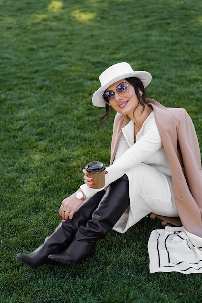 Fashion model with cup of coffee seat on the grass. Attractive woman weared whihe hat, dress, jacket and sunglasses.