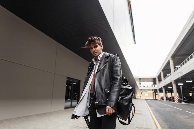 Fashion model young man in a stylish leather black jacket in a shirt in jeans with a fashionable backpack with a hairstyle posing near the road in the city. American guy hipster stands in the street.