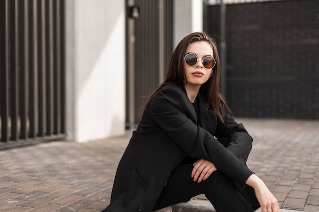 Fashion model young woman with sexy lips in stylish sunglasses in fashionable black clothes from new youth trendy collection is resting near building in city. Cool gorgeous girl hipster sit outdoors.