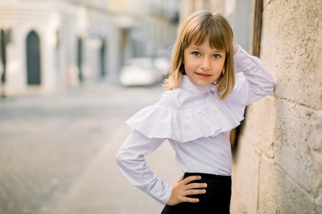 Photo fashion outdoor portrait of charming little girl in white blouse and black skirt, posing leaning on the wall of beautiful old city building in ancient european city. horizontal shot, copy space