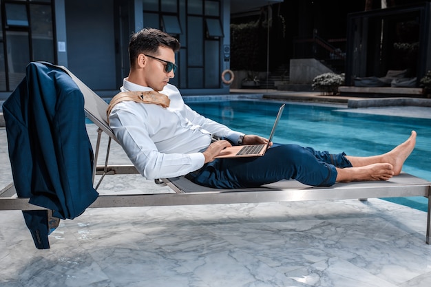 Fashion photo of smart man with a laptop relaxing on sun lounger near pool