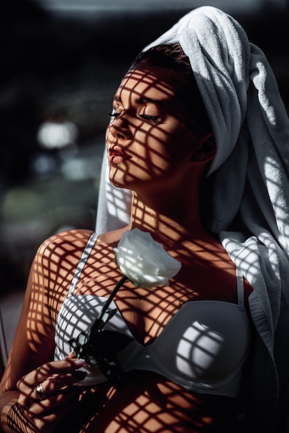 Fashion photo of young beautiful woman with closed eyes and shadows on her face. Brunette with a towel on her head, in white lingerie and a white flower in her hand, poses for the camera.