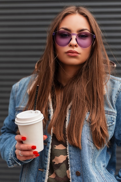 Fashion portrait American young woman hipster in stylish youth purple glasses in fashionable blue denim jacket with a hot drink in hands near gray wall on the street. Beautiful girl model in the city.