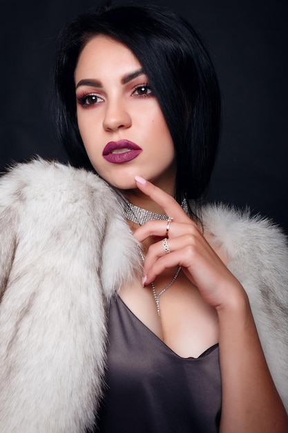 Fashion portrait of a beautiful brunette girl on fur with luxury accessories. Beauty Model with jewelery on black background. Girl in white Mink Fur Coat. Beautiful Luxury Winter Woman.