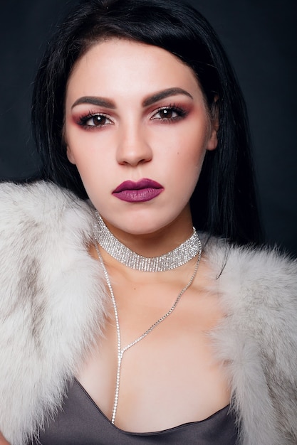 Fashion portrait of a beautiful brunette girl on fur with luxury accessories. Beauty Model with jewelery on black background. Girl in white Mink Fur Coat. Beautiful Luxury Winter Woman.