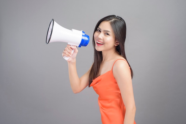 A fashion portrait of beautiful woman wearing orange dress is using megaphone isolated over gray