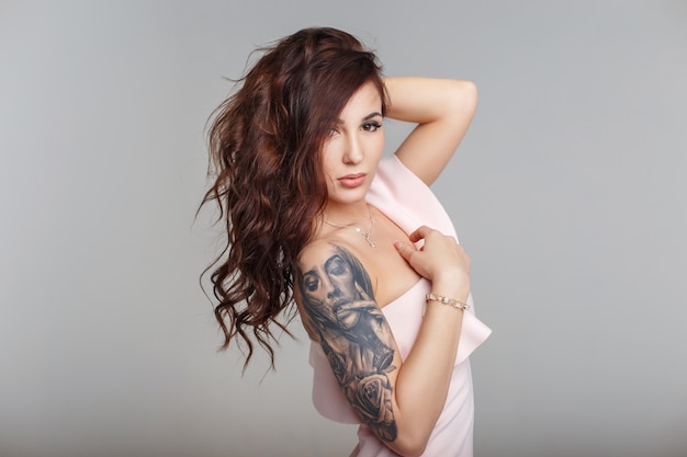 Fashion portrait of a beautiful young girl with a haircut with a tattoo on her arm in a stylish pink dress on a gray background