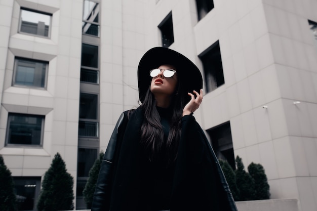 Photo fashion portrait of elegant young model dressed in black coat, stylish hat and glasses, posing near mall