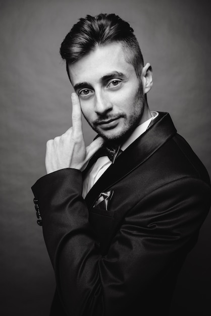 Fashion portrait of handsome elegant man with curly hair wearing tuxedo posing on gray background in studio. Black and white photo