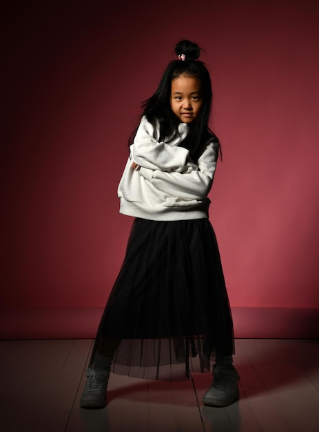 Fashion portrait of a little Korean model girl posing professionally on a red background Girl stands with folded arms and is wearing a black skirt white sweater sneakers Kids fashion concept