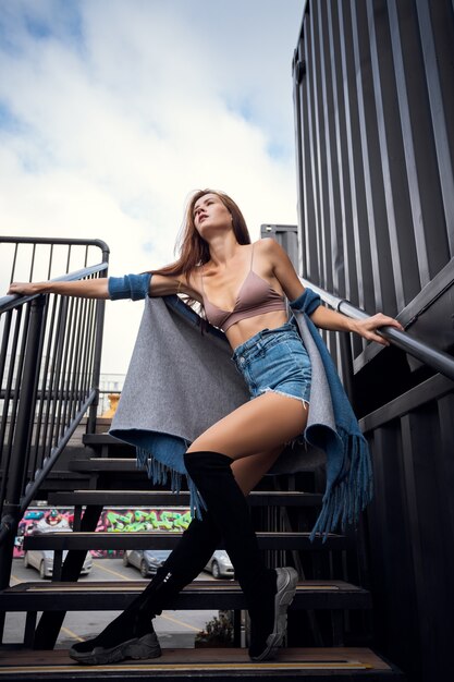 Fashion portrait of a young fashionable woman, dressed in sports and top, posing on the stairs, enjoys rest in a city, summer street fashion. portrait of a joyful woman