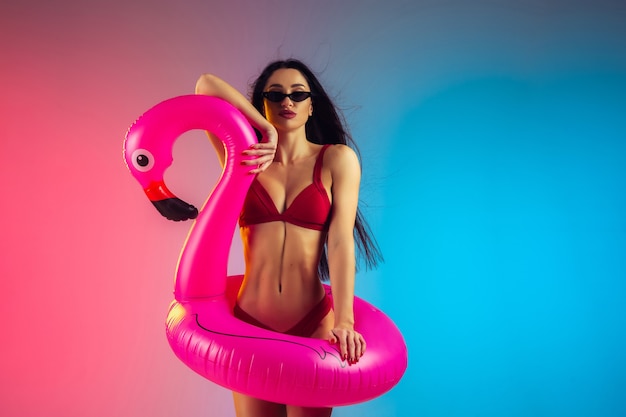 Fashion portrait of young fit and sportive woman with rubber flamingo in stylish red swimwear on gradient wall perfect body ready for summertime