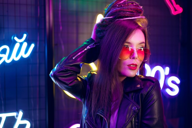 Fashion portrait of a young woman in sunglasses posing near neon signs in night club