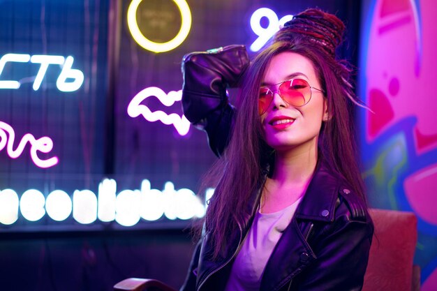 Fashion portrait of a young woman in sunglasses posing near neon signs in night club