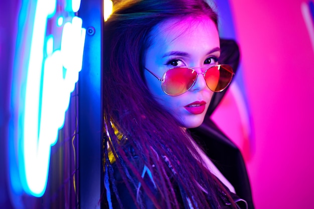 Fashion portrait of a young woman in sunglasses posing near neon signs in night club