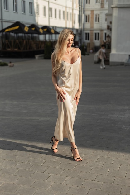 Fashion pretty elegance woman blonde model in stylish beige vintage long dress with shoes walks on the street at sunset light