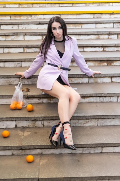 Fashion pretty woman model with fruit oranges sits on the steps in a purple jacket black shoes