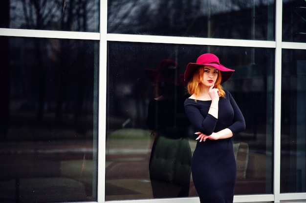 Fashion red haired girl on pink hat and black dress with bright make on large window