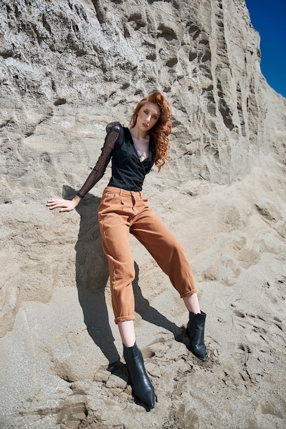 Fashion red-Haired woman in orange jeans posing in nature near sandy rocks