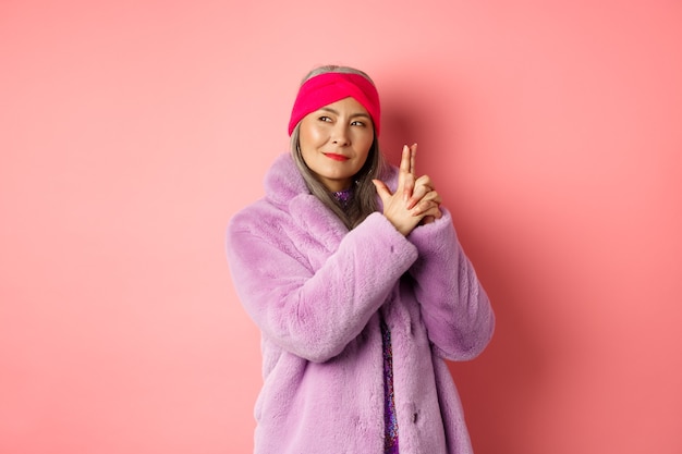 Fashion and shopping concept. Cool and stylish asian senior lady in purple fake fur coat, making finger gun gesture and looking left with sassy smile, acting like secret agent, pink background