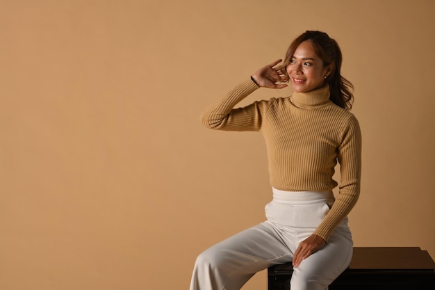 Fashion studio photo of beautiful lady in stylish warm sweater sitting on beige background Copy space for advertise text