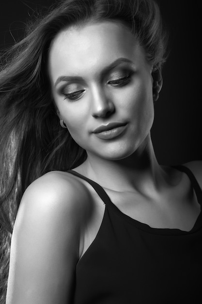 Fashion studio portrait of a luxurious brunette woman with perfect makeup and hairstyle wears classic black dress posing in a dark studio. Black and white shot