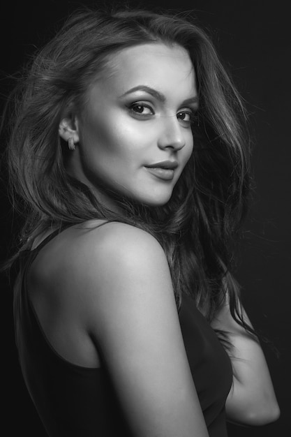 Fashion studio portrait of a wonderful brunette woman with perfect makeup and hairstyle wears classic black dress posing in a dark studio. Black and white shot