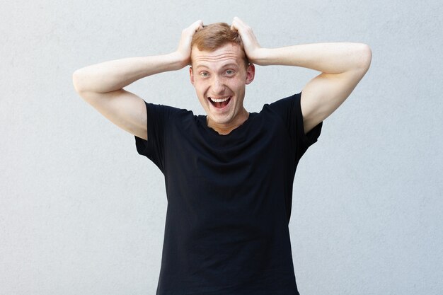 Fashion, style, emotions and people concept - Close up portrait of a redhead of a beautiful manly guy with freckles on a gray background black T-shirt. Holds his hands at the head and laughs