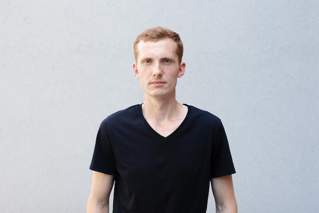 fashion style emotions and people concept Close up portrait of a redhead of a beautiful manly guy with freckles on a gray background black Tshirt looks at camera and smiles