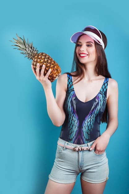 Fashion summer portrait beautiful woman in sun pink visor and pineapple over blue background