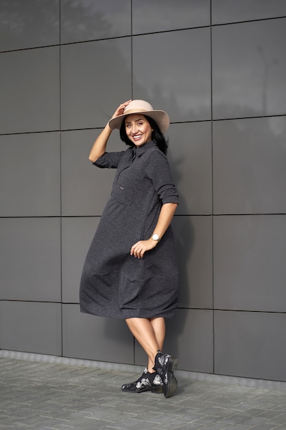 Fashion trendy collection. Happy funny beautiful brunette girl wearing stylish grey dress holding fashionable hat in windy day. Model in trendy outfit posing on grey wall