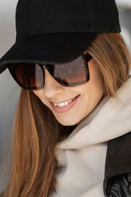 Fashion urban female portrait of beautiful trendy happy stylish woman hipster with smile with vintage sunglasses with black mockup cap in beige hoodie walks in the city