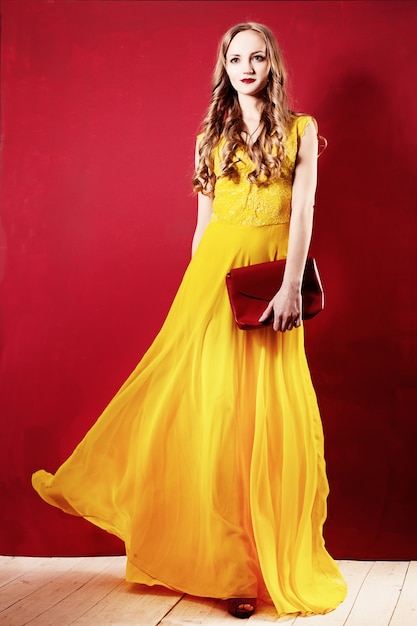 Fashion Woman in an Elegant Silk Dress. Curly Blonde Hair, Yellow Dress and Red wall