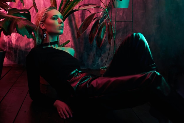 Fashion woman sitting on the floor in tropical foliage neon light Wet hair perfect figure and makeup Pink and green neon color Beautiful girl and monstera leaves