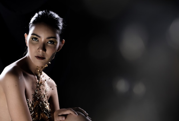 Fashion Young Asian Woman eyes black wrapped hair beautiful make up fashion decorate with Golden Foil or Gold leaf all over chest. Studio Lighting dark Background