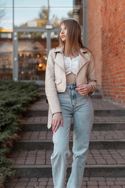 Fashion young beautiful girl Caucasian model in a fashionable white leather jacket with a top and vintage high waist jeans walks down the steps near a red brick mall