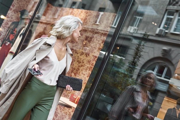 Fashion young stylish woman walking on the city street holding smartphone looking at reflection at