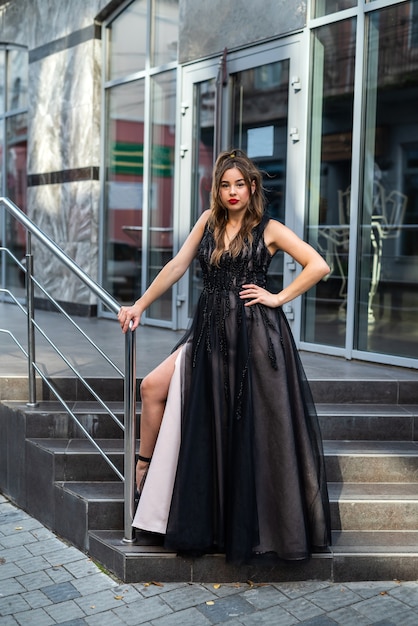 Fashion young woman full length in evening black dress walking in city daytime. Street style