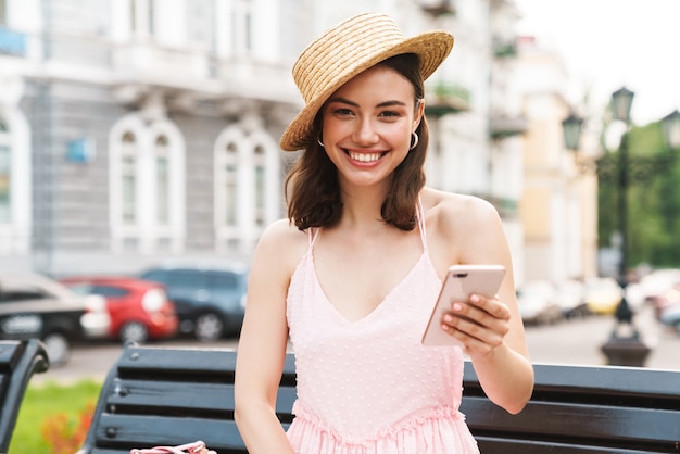 fashion young woman wearing summer straw hat smiling and holding cellphone while walking on city street