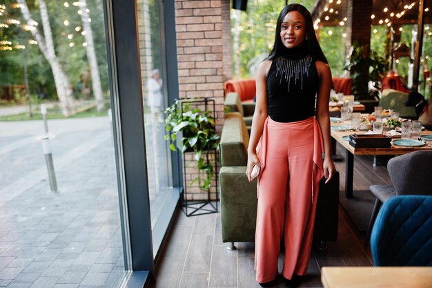 Fashionable african american woman in peach pants and black blouse pose at restaurant.